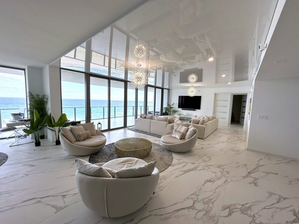 The same oceanfront living room with a gloss stretch ceiling reflecting the water, recessed downlights and a perimeter cove, after